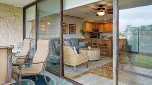 A spacious living area with a glass sliding door leading to a kitchen and dining area, featuring wicker furniture and a ceiling fan. - 2 bedroom updated Kihei condo, AC, walk to beach, shops, & dining (Kihei)