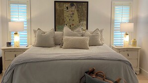 Un dormitorio con cama, ventilador de techo, un cuadro, mesitas de noche con lámparas y un banco.