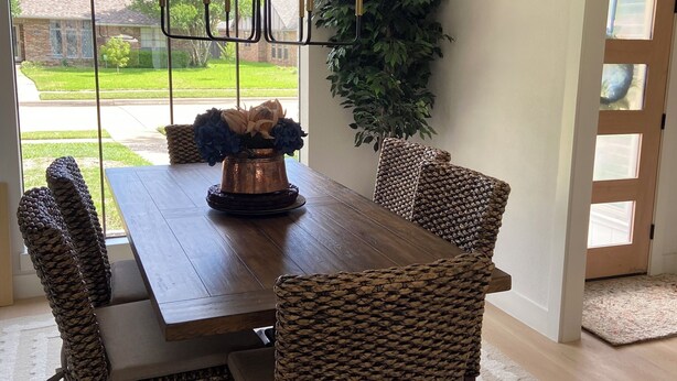 Un comedor con mesa de madera, sillas de mimbre, candelabro y ventana grande con vista a una casa y jardín.