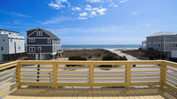 A wooden deck with a railing overlooking a beach with houses and a clear sky. - Charming 4-Bedroom Home with Ocean Views in Topsail Beach! (Topsail Beach)