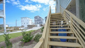 A wooden staircase leading to a building with multiple floors, a clear sky, and a landscaped area with shrubs. - Charming 4-Bedroom Home with Ocean Views in Topsail Beach! (Topsail Beach)