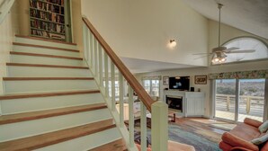 A staircase with wooden steps and white railings leading to a living area with a fireplace, a television, and a large window with a view. - Charming 4-Bedroom Home with Ocean Views in Topsail Beach! (Topsail Beach)