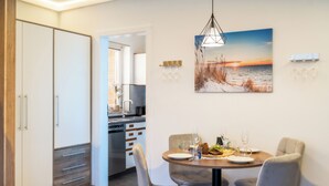 Een eetgedeelte met een tafel gedekt voor twee personen, een keuken op de achtergrond en een schilderij van een ondergaande zon aan de muur.