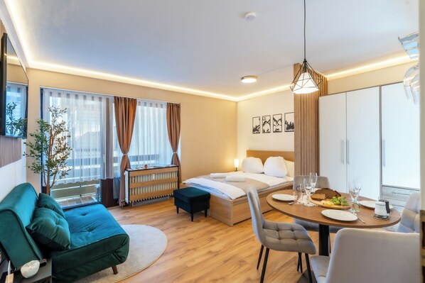 Quarto de hotel moderno com cama, mesa de jantar com cadeiras, sofá, radiador e janela com cortinas.