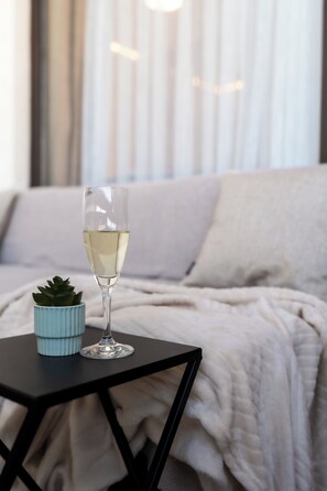 Um copo de vinho branco sobre uma mesa pequena, ao lado de um vaso com planta, com uma cama e cortinas ao fundo.