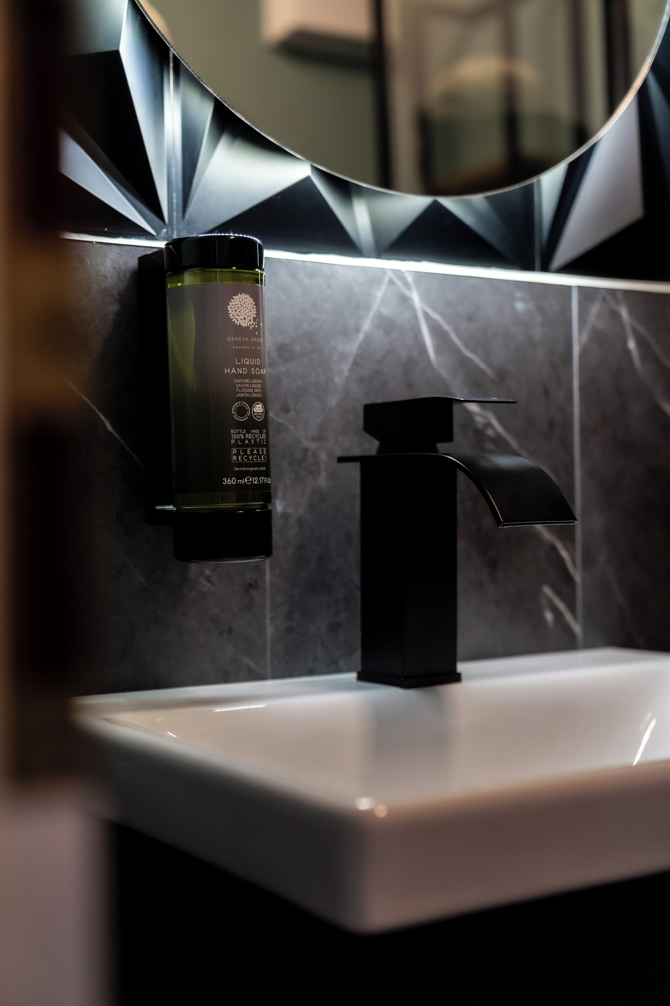 A bathroom with a modern sink, a black faucet, and a bottle of liquid soap on the wall.