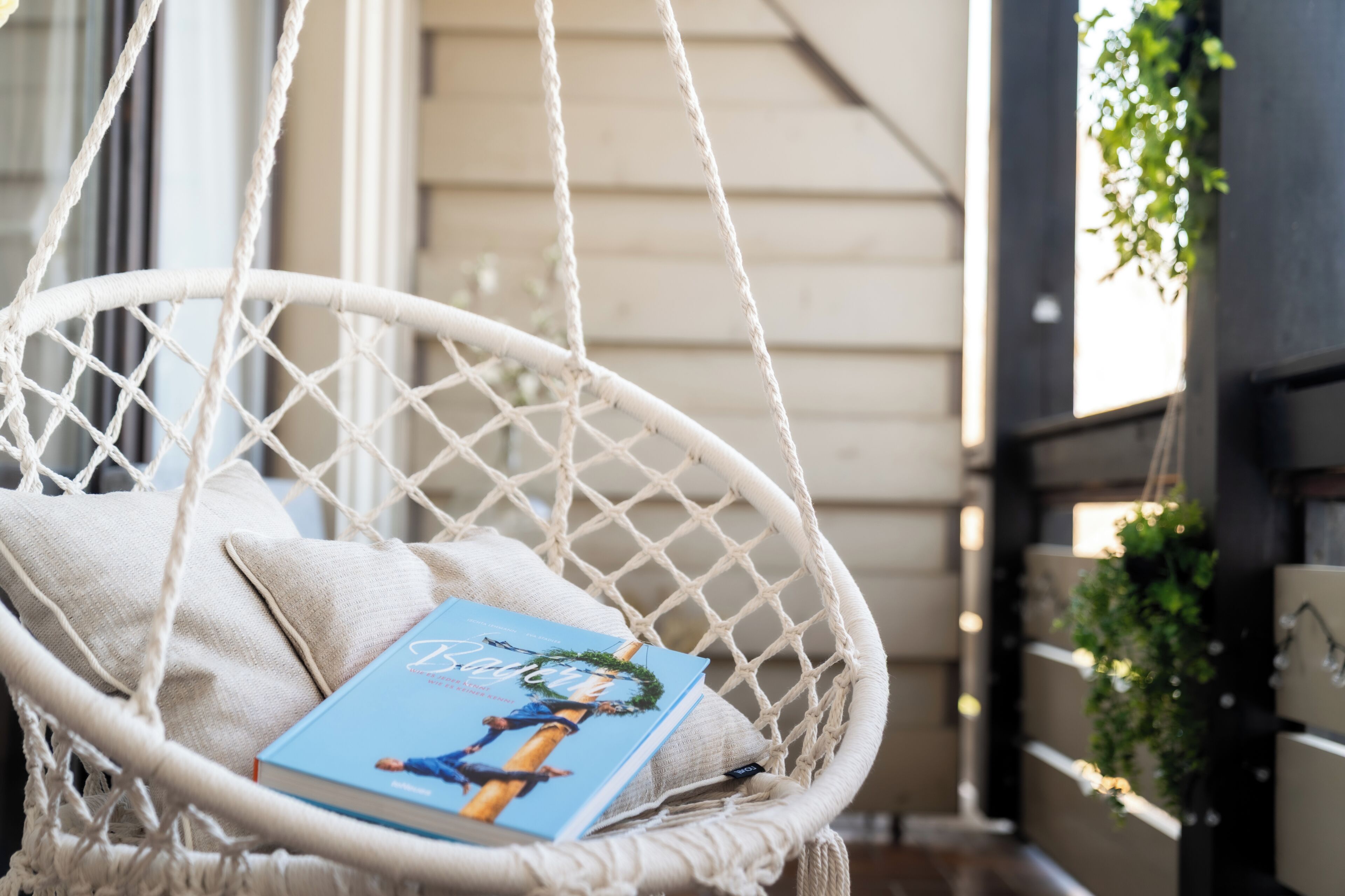 Een witte rieten stoel met een boek en kussen, een houten wand en een hangplant.