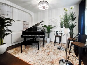 A piano, chairs, a table, and potted plants in a room with a patterned rug and a chandelier.