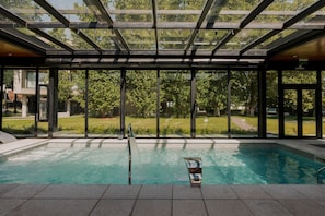 Piscina com teto de vidro, rodeada por vista para o jardim.