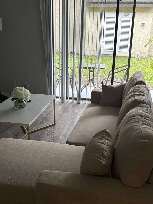 A modern living room with a sofa, a dining table with a vase of flowers, and a view of a patio with a table and chairs.