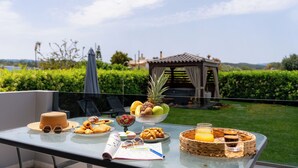 Een tafel met eten en drinken, een hoed, zonnebril en een boek op een balkon met uitzicht op een tuin en een tuinhuis.