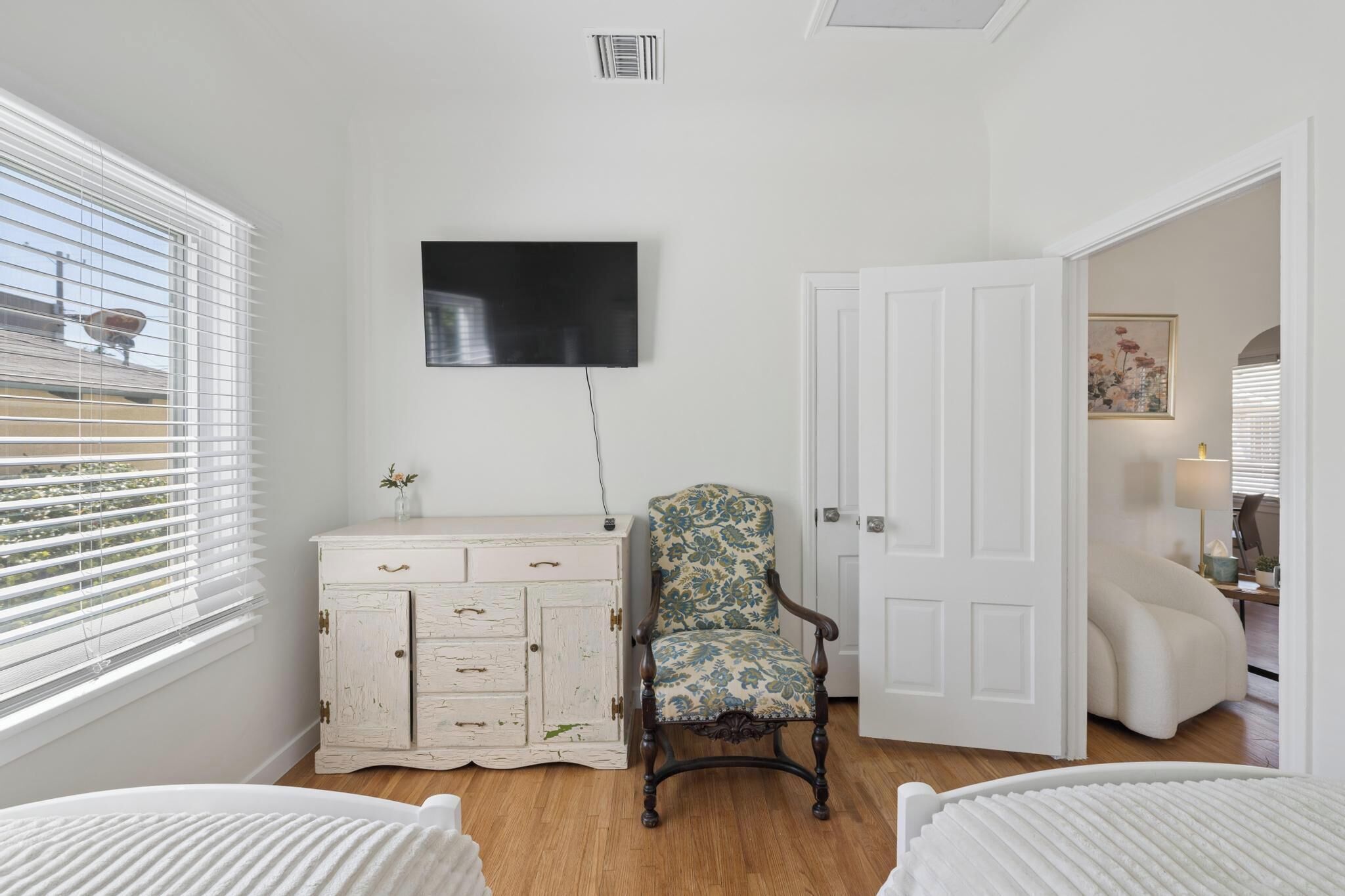 Une pièce avec une commode blanche, un fauteuil à motifs floraux, un téléviseur à écran plat et une porte donnant sur une autre pièce.