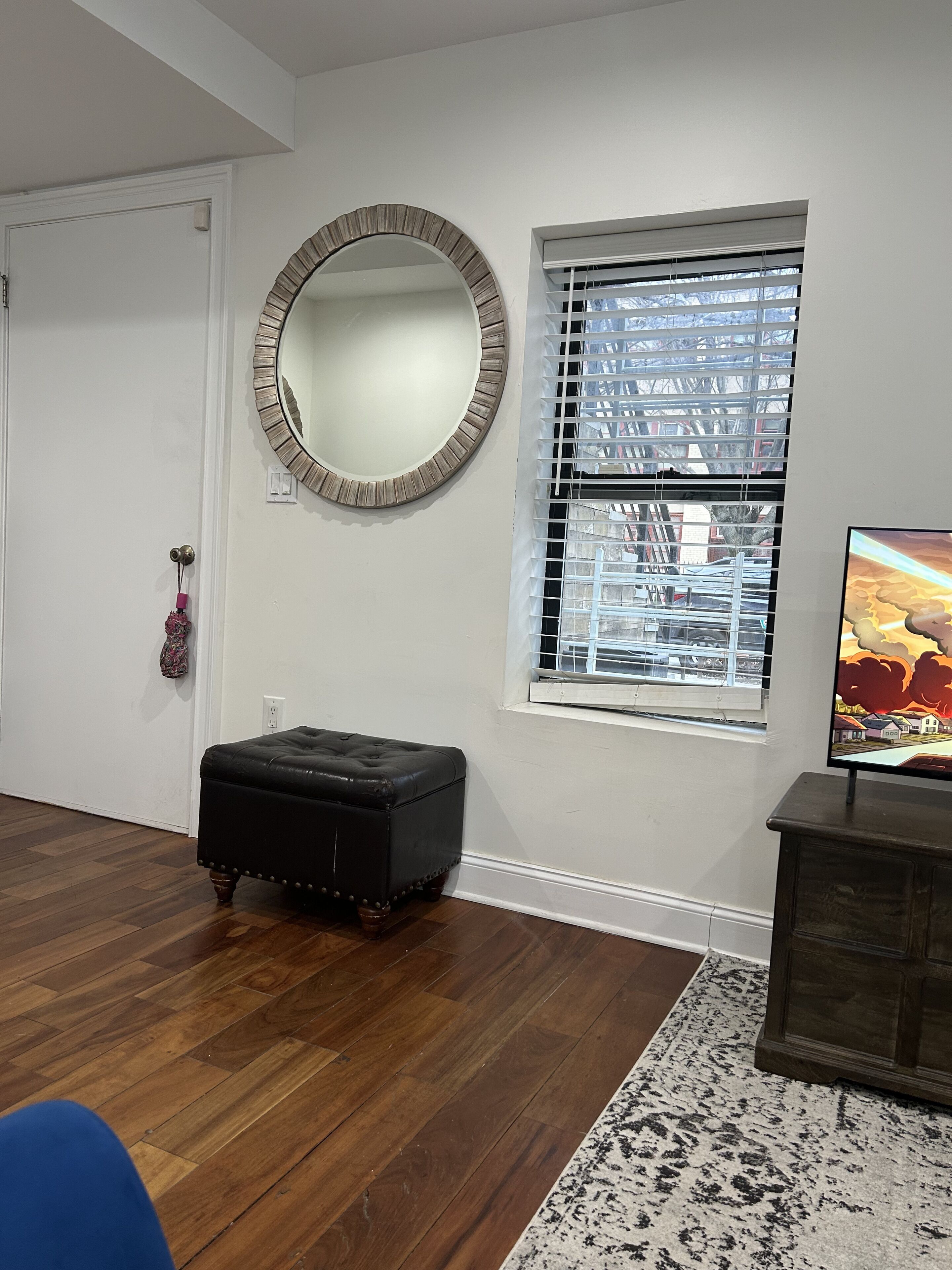 Une pièce avec un plancher en bois, un miroir rond, une fenêtre avec des stores, un pouf noir et un téléviseur posé sur un meuble en bois.