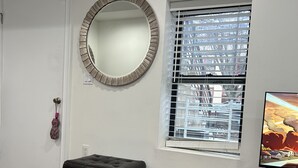 Een kamer met houten vloer, een ronde spiegel, een raam met zonwering, een zwart voetenbankje en een televisie op een houten dressoir.