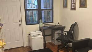 Een kamer met een bureau, stoel en een zwart tapijt. Er is een raam met glas-in-lood, een witte deur en ingelijste foto's aan de muur.