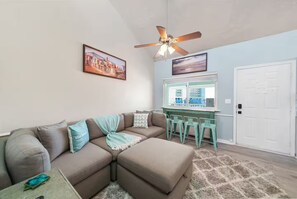 A living room with a sectional sofa, a ceiling fan, and a window with a view of a building. - West End, Beautiful Ocean Views, Across From Beach, Pool (Panama City Beach)