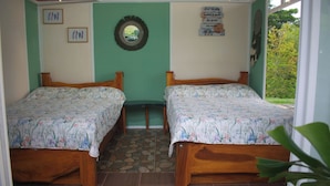 A room with two wooden beds, a stone-tiled floor, a green wall, and a view of greenery outside.