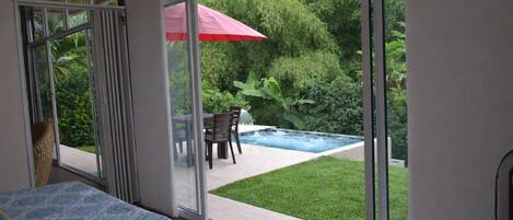A room with a bed, a large sliding glass door leading to an outdoor area with a pool, patio furniture, and lush greenery.