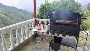 A barbecue grill on a balcony with a scenic view of hills, a table with condiments, and a metal chair. - El Mirador De Serrato (La Vega)