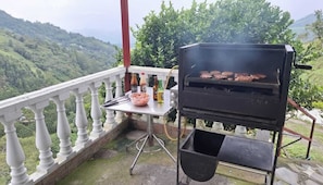 A barbecue grill on a balcony with a scenic view of hills, a table with condiments, and a metal chair.