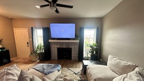 Una sala de estar con televisor de pantalla plana, chimenea, ventilador de techo y dos ventanas con cortinas.