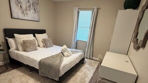 Un dormitorio con cama, escritorio, ventana con cortinas, luz de techo y un cuadro en la pared.