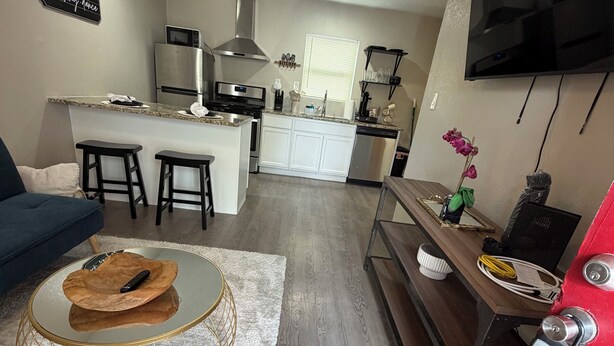 Una cocina moderna con microondas, estufa y lavabo, un comedor con mesa redonda y taburetes, y una sala de estar con sofá y televisor montado en la pared.