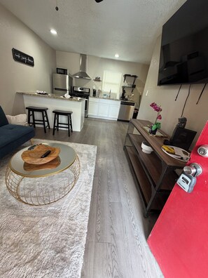 Una cocina moderna con microondas, estufa y lavabo, un comedor con mesa redonda y taburetes, y una sala de estar con sofá y televisor montado en la pared.