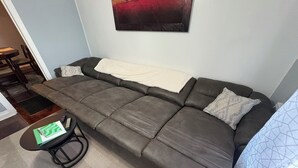 Um sofá cinzento com um cobertor branco, uma pequena mesa de apoio com um caderno verde e um quadro na parede.