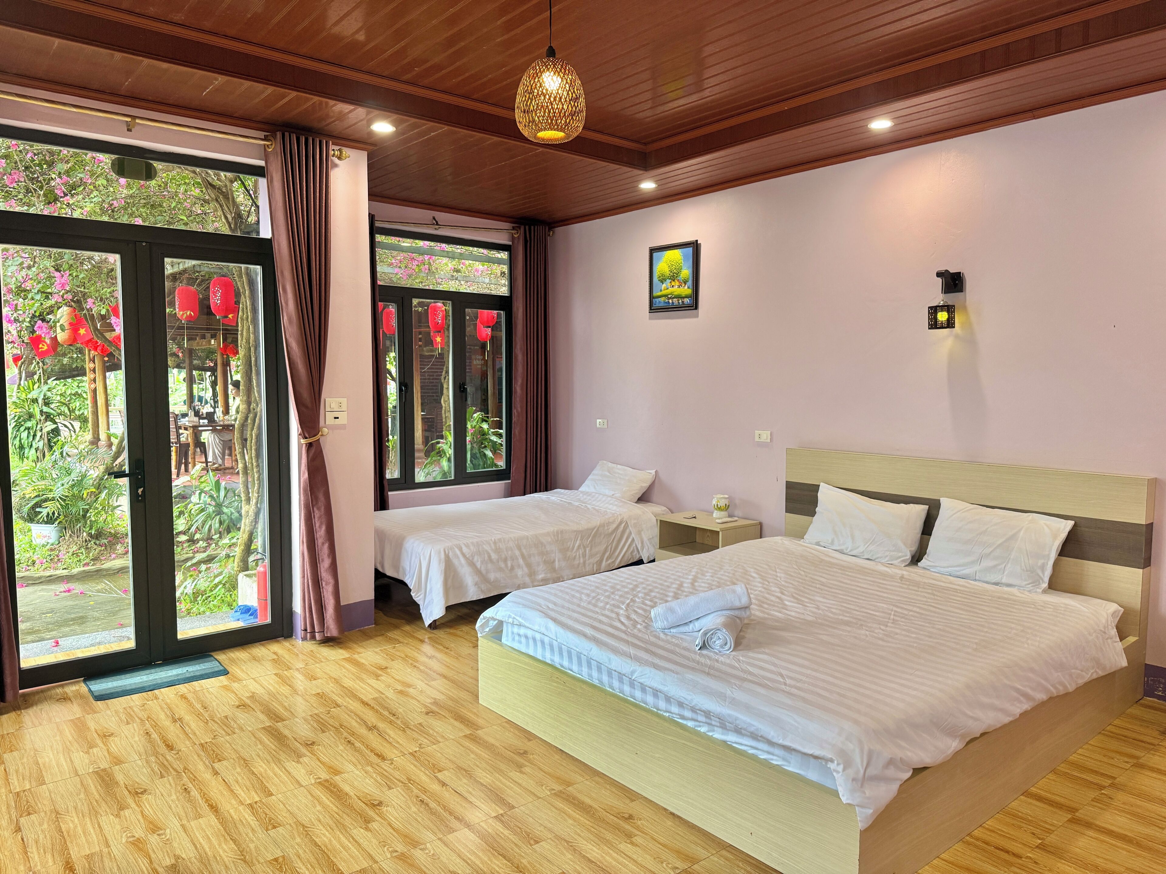 A hotel room with two beds, a wooden floor, a large window with curtains, a hanging light fixture, and a small table with a lamp.