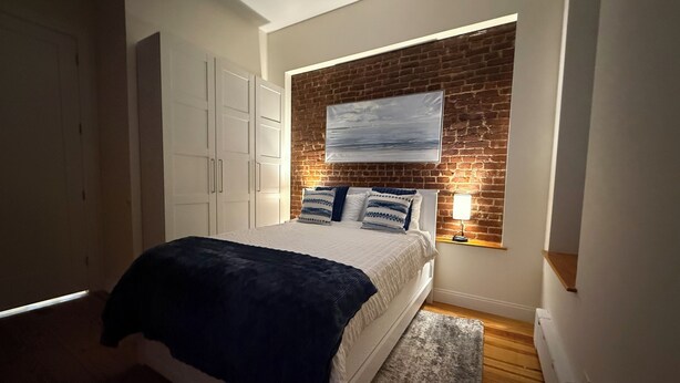 Un dormitorio con cama, mesita de noche, lámpara, ventana con vistas y pared de ladrillo.