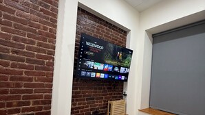 Una sala de estar moderna con pared de ladrillo, un televisor montado mostrando una película, una pantalla de proyector y un área de descanso acogedora con cojines.