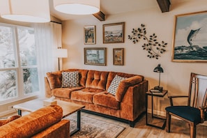 Ein Wohnzimmer mit einem braunen Sofa, einem Couchtisch, einem Beistelltisch mit Lampe und einem Sessel. An der Wand befinden sich Bilder in Rahmen und ein Fenster mit Vorhängen.