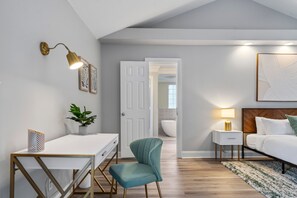 Ein Schlafzimmer mit Bett, Nachttisch, Lampe, Stuhl, Schreibtisch und einer Tür zu einem Badezimmer.