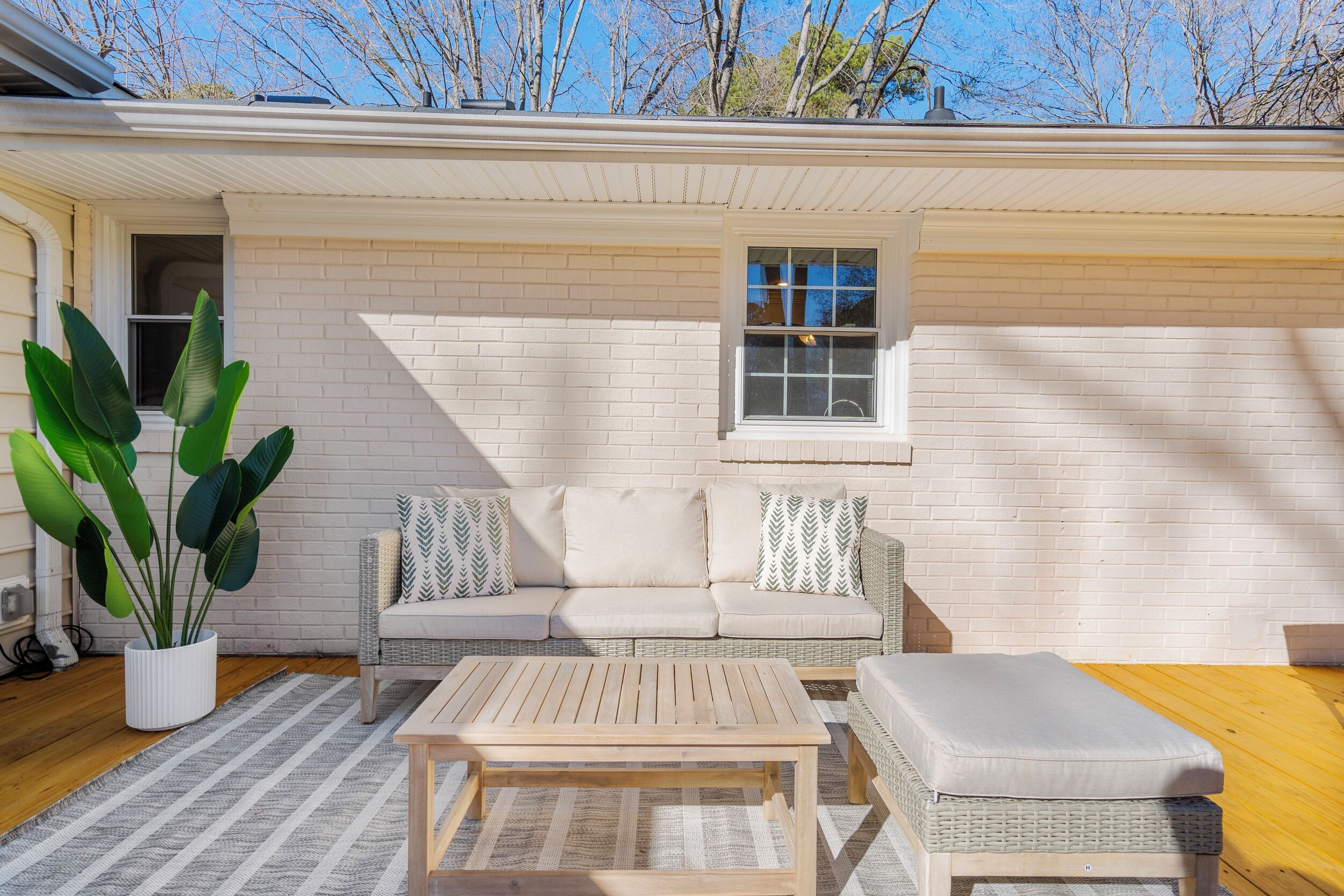 A cozy outdoor seating area with a white brick wall, a wicker sofa, a wooden table, and a large potted plant.