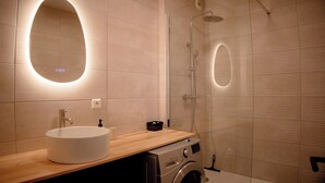 Uma casa de banho moderna com um espelho oval grande, uma pia, uma máquina de lavar roupa e um chuveiro.