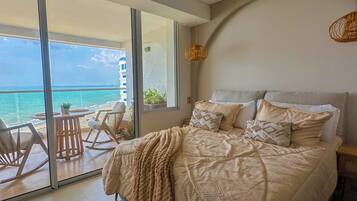 A bedroom with a large bed, a ceiling fan, a view of the ocean, and a balcony with chairs.