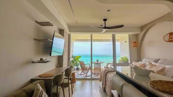 A modern living room with a sofa, a dining table, and a TV. There is a balcony with a view of the sea.
