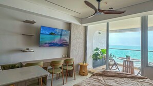 Quarto moderno com cama grande, televisão, escrivaninha e cadeiras, e varanda com vista para o mar.