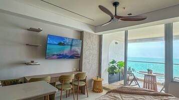 A modern bedroom with a large bed, a TV, a desk, and chairs, and a balcony with a view of the sea.