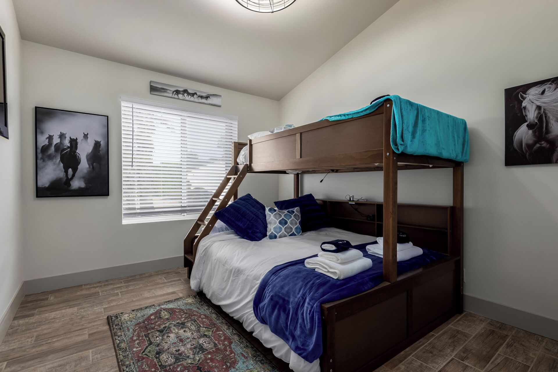 Un lit superposé avec une structure en bois et une couverture bleue, un tapis à motifs et une fenêtre avec des stores.