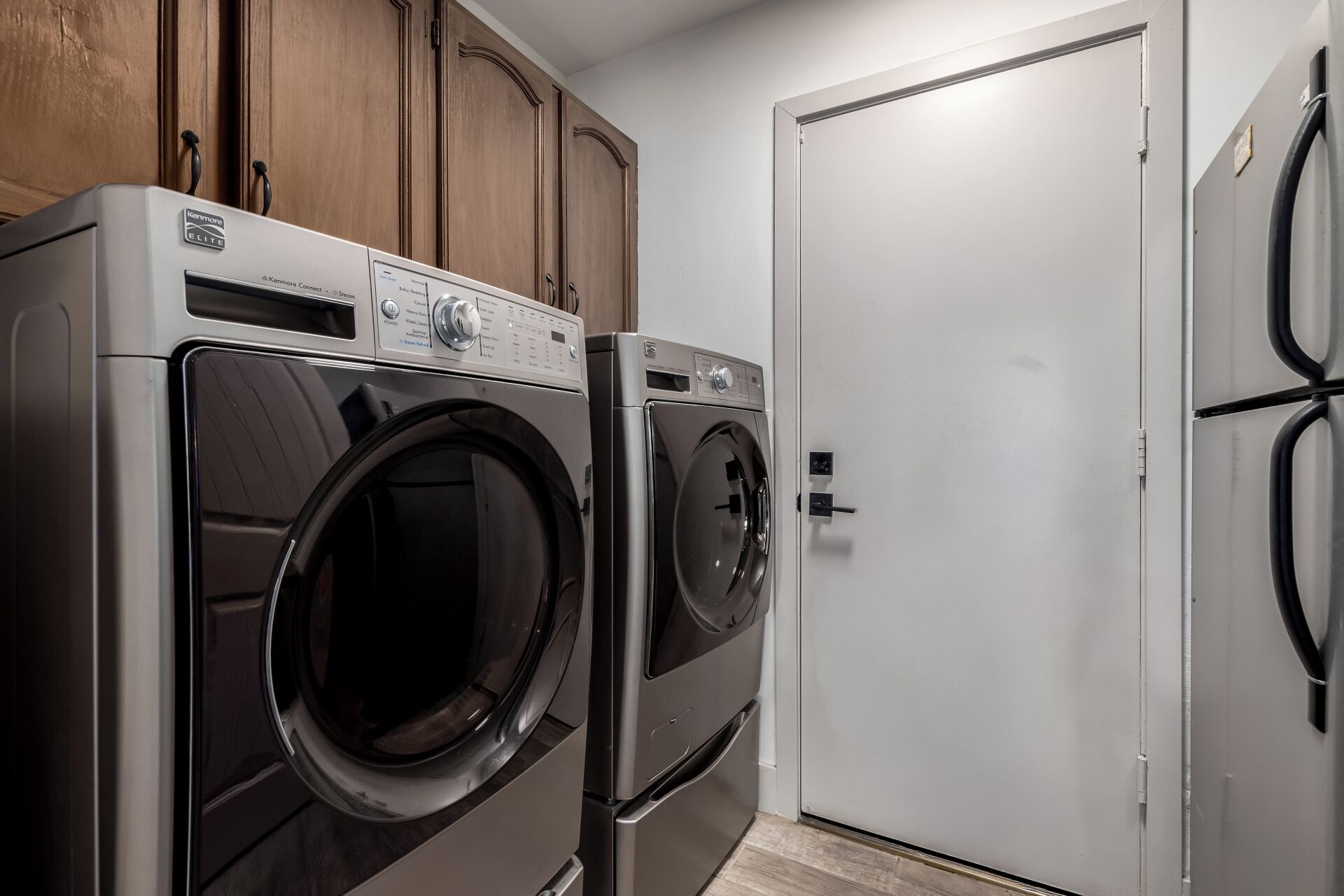 A compact laundry room with two Kenmore Elite front-loading washers, wooden cabinets above, and a white door.