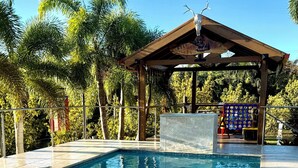 A swimming pool with a wooden deck, surrounded by palm trees and a covered area with a sign.