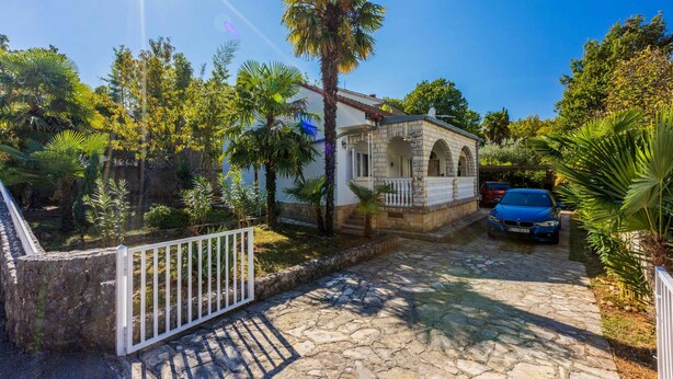 A stone pathway leading to a building with a white fence and a blue car parked nearby. - Holiday apartment with terrace and barbecue (Malinska)