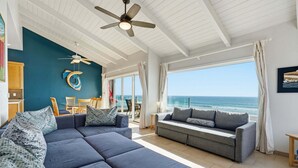 A spacious living area with a large sofa, a smaller couch, and a ceiling fan. There is a view of the ocean through large windows. - Oceanfront Condo | Balcony, Fire Pit | Shared Beach | Sleeps 6 (Oceanside)