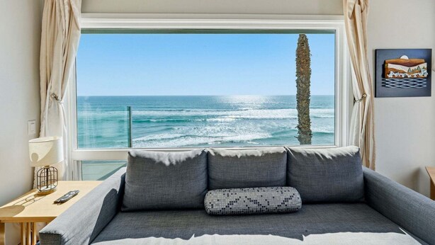 A bedroom with a large window overlooking the ocean, a bed with grey bedding, a lamp, and a remote control on a wooden nightstand. - Oceanfront Condo | Balcony, Fire Pit | Shared Beach | Sleeps 6 (Oceanside)