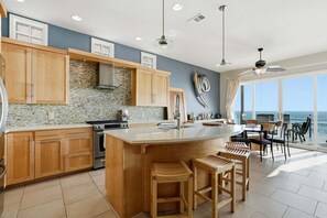 A modern kitchen with wooden cabinets, a large island, and a view of the ocean. - Oceanfront 3BR Condo | Balcony, Fire Pit | Shared Beach | Sleeps 6 (Oceanside)