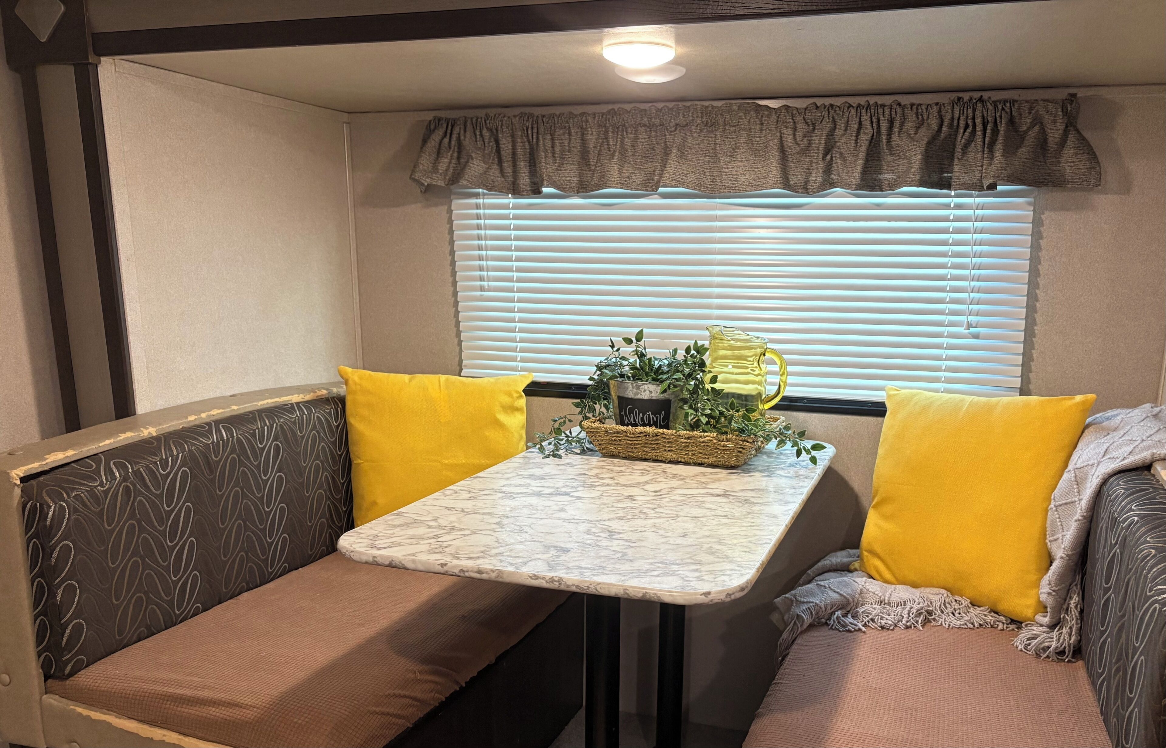 Interior compacto de una autocaravana, con asientos acolchados, una mesa pequeña y una cesta de plantas, además de una ventana con persianas.