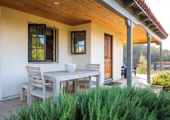 A covered patio with a wooden table and chairs, a large window, and a door leading to an outdoor area. - Modern Farmhouse in the Hills (Goleta)