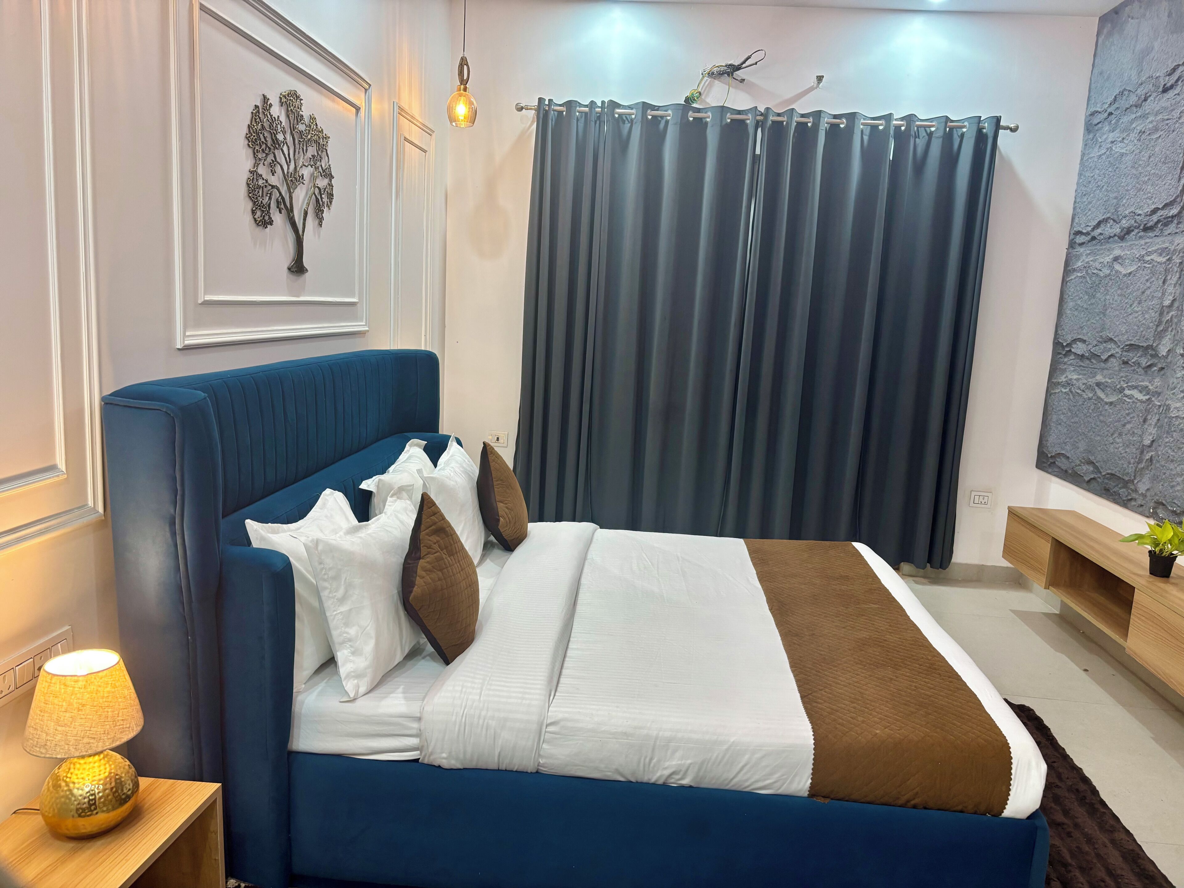 A bedroom with a blue bed, white and brown pillows, a bedside lamp, a wall-mounted artwork, and a curtain.